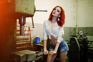 Red haired girl wear on short denim shorts and white blouse posed at industrial machine at the factory. photo