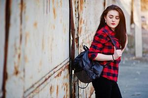 Fashion portrait girl with red lips wearing a red checkered shirt and backpack background rusty fence. photo