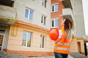 Back of engineer builder woman in uniform waistcoat and orange protective helmet against new building. Property living block theme. photo