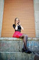 Portrait of girl with black choker on her neck, red leather skirt and mobile phone at hand with headphones against orange shutter. photo