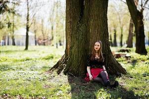 Portrait of girl with bright make up with red lips, black choker necklace on her neck and red leather skirt sitting near tree at park. photo