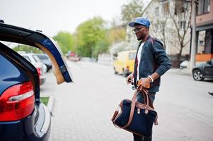 retrato de hombre afroamericano con estilo en ropa deportiva, gorra y gafas caminando con bolso y maletero abierto. los hombres negros modelan la moda callejera. foto
