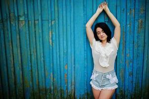 Portrait of sexy brunette girl on women's jeans shorts and white blouse against blue wooden background. photo