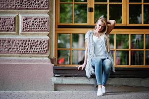 Elegante mujer rubia vestida con jeans y una chica sin mangas con camisa blanca contra las ventanas de la calle. retrato de modelo urbano de moda. foto