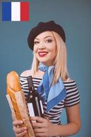 portrait of beautiful blonde french woman in beret, scarf, back and white shirt, with bottle of wine and bread baguette in her arms with French flag on background photo
