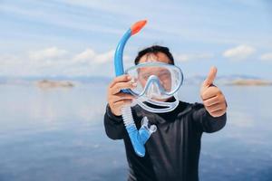 Asian man in wetsuit holding snorkeling mask and giving thumb up on summer vacation