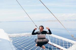 Excited Asian man in wetsuit and headphone sitting on catamaran net and raising his hand with excited expressing winning gesture while looking in a laptop on vacation at Labuan Bajo photo