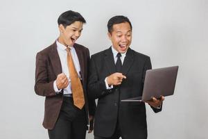 dos felices hombres de negocios asiáticos en traje usando una computadora portátil con expresión conmocionada y gesto ganador foto