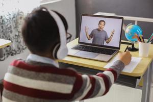 A schoolboy is studying and watching his teacher on a laptop while writing on a paper photo