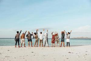 vista trasera de un grupo de amigos de pie y levantando la mano juntos disfrutando de la vista al mar desde la playa durante las vacaciones de verano foto