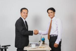 Two asian businessmen wearing suits and ties shaking hands while looking to camera between office desks for deal concept photo