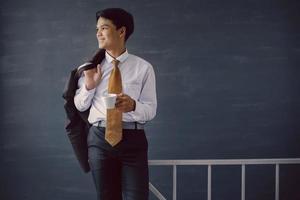 hombre de negocios asiático con camisa y corbata sonriendo sosteniendo un café y una chaqueta de traje en el hombro foto