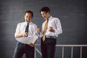 Happy two Asian businessman shocked and surprised looking to paper of report with winning gesture photo
