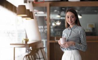 mujer de negocios asiática sonriente sosteniendo una taza de café y una computadora portátil en la oficina. mirando a la cámara. foto