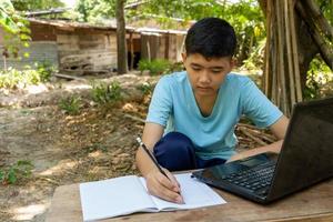 A boy is writing in a notebook while studying online with a laptop computer at home in the countryside photo