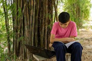 A boy sits in a wooden chair studying online with a laptop computer in a bamboo tree in the countryside photo