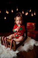 Una niña de 7 meses con un traje rojo de Navidad sobre un fondo de guirnaldas retro se sienta en una piel foto