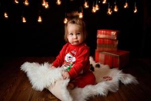 Una niña de 7 meses con un traje rojo de Navidad sobre un fondo de guirnaldas retro se sienta en una piel foto