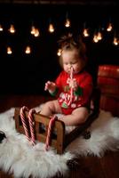 Una niña de 7 meses con un traje rojo de Navidad sobre un fondo de guirnaldas retro se sienta en una piel foto