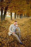 Portrait of a young man in a coat on a background of autumn trees. A man sits on the ground. photo