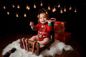 Una niña de 7 meses con un traje rojo de Navidad sobre un fondo de guirnaldas retro se sienta en una piel foto