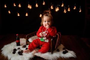 Una niña de 7 meses con un traje rojo de Navidad sobre un fondo de guirnaldas retro se sienta en una piel foto