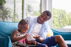 Cheerful african american father and daughter playing in living room, Cute little girl sitting on the sofa and playing on tablet photo