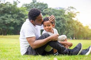 feliz padre afroamericano mientras abrazaba y cargaba a su hijo, papá estaba besando a su hijo en el parque, alegre concepto de familia negra. foto