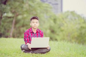 Boy sitting on the grass studying with laptop in the park, looking at camera , Outdoor learning concept photo