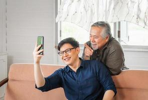 Cheerful senior asian father and adult son using smart phone talking on video call in living room photo