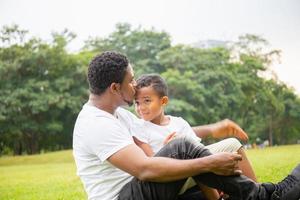 feliz padre afroamericano mientras abrazaba y cargaba a su hijo, papá estaba besando a su hijo en el parque, alegre concepto de familia negra. foto