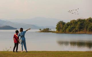 mother and son standing beside big lake and see mountain view in the background, mom pointing finger to birds flying in sky. Idea for family tourist travels together