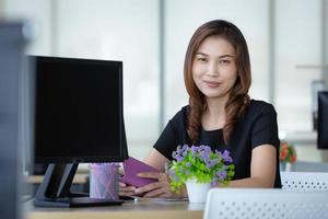 mujer de negocios asiática senior sentada en la oficina cerca de la pantalla de la computadora y mirando a la cámara con confianza en sí misma foto