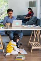 familia de raza mixta compartiendo tiempo en la sala de estar. padre caucásico usando una computadora portátil para trabajar y medio tailandés jugando y pintando debajo del escritorio mientras una madre asiática con una computadora portátil trabaja en su trabajo en el sofá foto