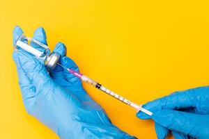 Hands in blue gloves holding syringe and stabbing to a bottle of a vaccine on orange background with copy space photo