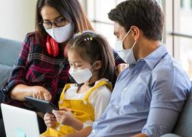 Mixed race family stays at home together with wearing protective hygiene mask on faces, father and mother helping and teaching little girl to use tablet computer in learning photo