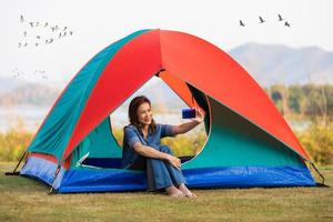 A beautiful woman sitting in a camping tent and using smartphone taking selfie for herself with big lake in background and group of birds flying in morning. photo