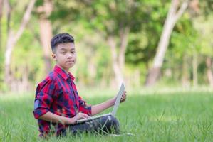 niño sentado en el césped estudiando con una laptop en el parque, mirando la cámara, concepto de aprendizaje al aire libre