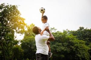 alegre padre afroamericano e hijo jugando al fútbol en el parque, conceptos familiares de felicidad foto