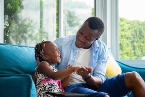 Cheerful african american father and daughter playing in living room, Cute little girl sitting on the sofa and playing on tablet photo