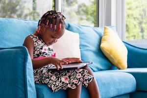 Cute little african american girl sitting on the sofa and playing on tablet in the living room photo