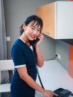Asian woman chambermaid in uniform speaking on room telephone during work. She smiles and looking at the camera takes orders from the hotel administrator.