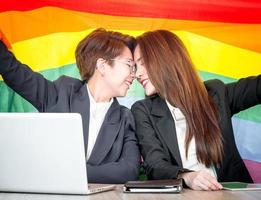Happy lesbian, beautiful Asian young two women, girl gay, couple love moment spending good time together, holding LGBT rainbow, pride flag on the working table together at the coffee shop. photo