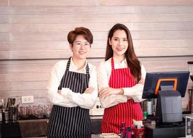 Young confident Asian lesbian couple who owns a coffee shop and a barista standing at the bar and waiting for customer orders from a computer in the coffee shop
