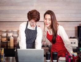 Young confident Asian lesbian couple who owns a coffee shop and a barista standing at the bar and checking online customer orders from a computer in the coffee shop