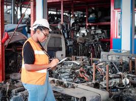 una mujer africana que trabaja en el almacén de repuestos automotrices usa un casco de seguridad y toma nota en el portapapeles entre los viejos motores de repuestos automotrices. foto