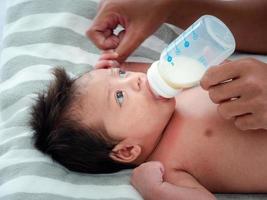 el retrato de una madre asiática alimenta al recién nacido asiático australiano de tres semanas con fórmula láctea de un biberón, concepto de maternidad e infancia o bebé acostado en la cama blanca. foto