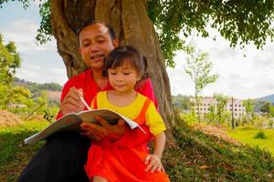 The father teaching daughter outside on filed under the tree. Father and daughter reading book washing hand under the tree. photo