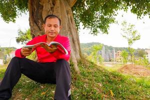 un joven leyendo un libro lavándose la mano afuera bajo el árbol. foto