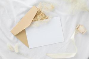 maqueta de invitación de tarjeta de felicitación en blanco 5x7 en sobre con flores secas y cinta sobre fondo de tela blanca, puesta plana, maqueta foto